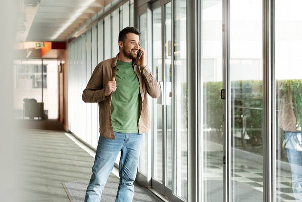 Красивый улыбающийся латиноамериканец, разговаривающий по мобильному телефону, стоящий в современном офисе и смотрящий в сторону, копирующий пространство. Технологическая концепция - Фото, изображение