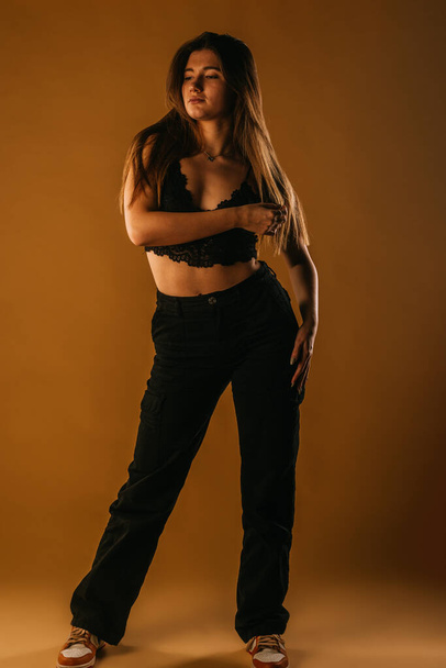 Young fashion girl dressed in black bra, black pants and sports sneakers standing and posing on a beige background in studio - Foto, Bild