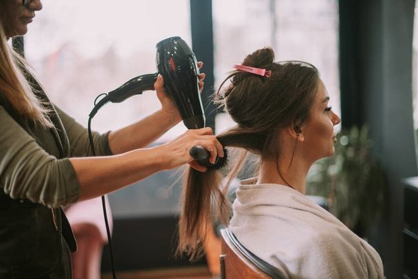 Peluquería rubia cepillando el pelo castaño liso de sus clientes mientras se sienta en el salón de belleza - Foto, Imagen