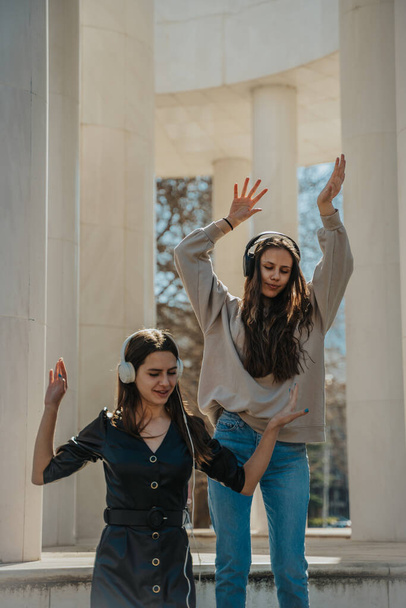 Two attractive brunette girls listening to music on their headphones and dancing with their hands up. They are hanging out in the city - Φωτογραφία, εικόνα