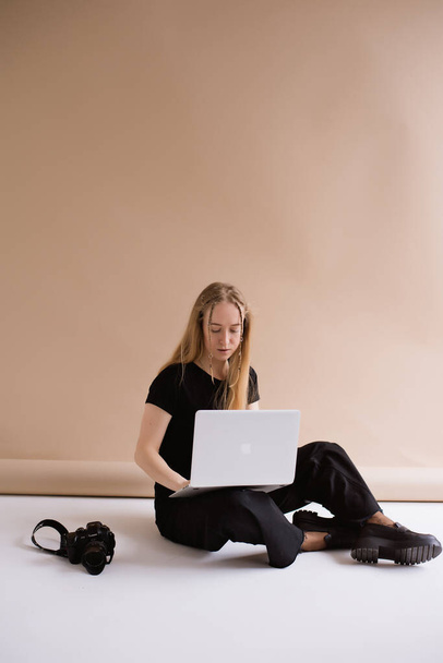 A business woman freelancer in a black is working, typing on a laptop apple and sitting on a white floor. blonde assistant in video production with computer MacBook and camera .  - Photo, Image