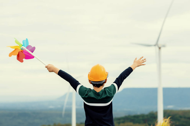 Progressive young asian boy playing with wind pinwheel toy in the wind turbine farm, green field over the hill. Green energy from renewable electric wind generator. Windmill in the countryside concept - Foto, Imagem