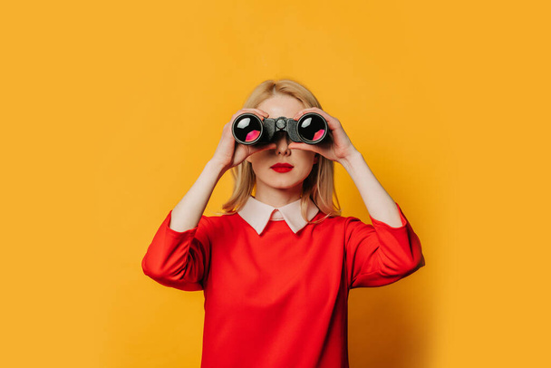 Stylish blonde hair woman in red dress with binoculars on yellow background - Foto, immagini
