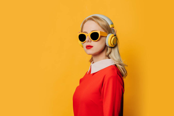 Stylish blonde hair woman in red dress with headphones on yellow background - Фото, изображение