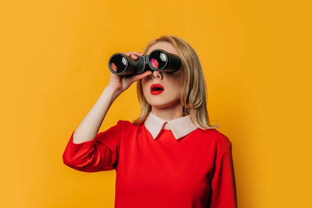 Surprised blonde hair woman in red dress with binoculars on yellow background - Fotografie, Obrázek