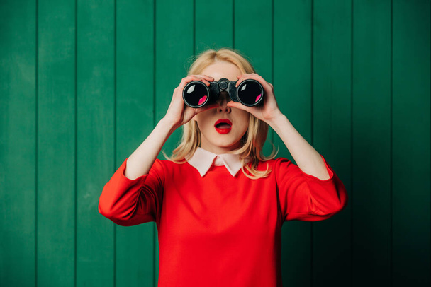 Surprised blonde hair woman in red dress with binoculars on green wooden background - Photo, Image