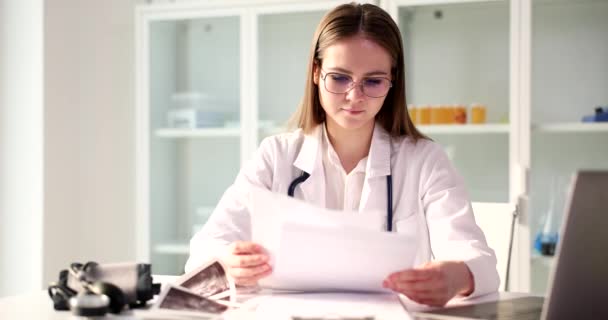 Ο εστιασμένος γιατρός εξετάζει τα υλικά των ασθενών που ψάχνουν στο laptop. Νεαρή γυναίκα με γυαλιά μελετά το ιατρικό ιστορικό κάθεται στο τραπέζι σε αργή κίνηση γραφείου - Πλάνα, βίντεο