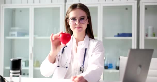 Cardiologue positif tient le modèle de coeur rouge dans la main regardant à la caméra. Jeune femme médecin dans des lunettes se trouve près d'un ordinateur portable ouvert souriant au ralenti - Séquence, vidéo