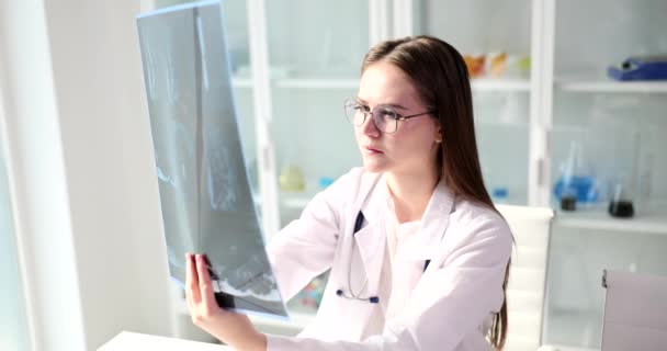 Концентрированный врач изучает рентгеновское изображение позвоночника пациента, сидящего за столом. Молодая женщина в очках ставит диагноз глядя на рентгенографию - Кадры, видео