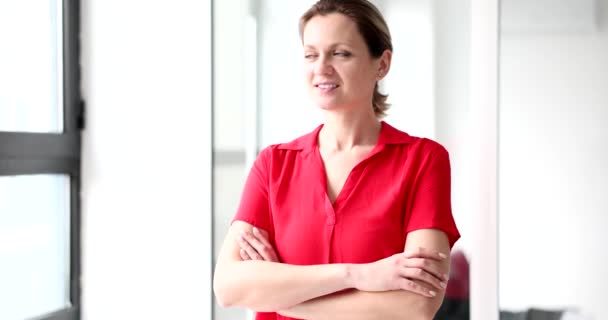 Θετική γυναίκα διευθυντής με κόκκινη μπλούζα γυρίζει να κοιτάξει έξω από το φωτεινό παράθυρο. Ευτυχισμένη γυναίκα στέκεται στο γραφείο με σταυρωμένα χέρια χαμογελώντας αργή κίνηση - Πλάνα, βίντεο