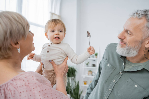 donna che tiene allegra nipote con cucchiaio vicino barbuto marito dai capelli grigi in cucina - Foto, immagini
