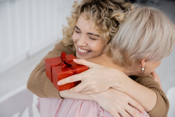 überglückliche Frau mit geschlossenen Augen hält Geschenkbox und umarmt Mutter mittleren Alters zu Hause - Foto, Bild