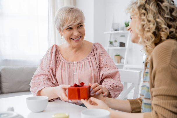 pleased middle aged woman holding present and looking at adult daughter while sitting near tea cups in kitchen - Fotoğraf, Görsel