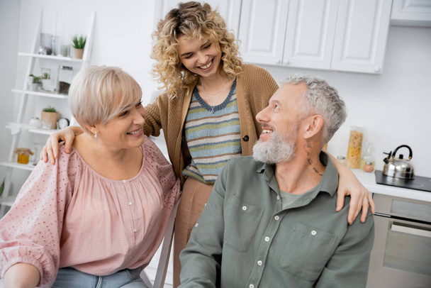 cheerful woman with curly blonde hair hugging happy parents in kitchen - Fotoğraf, Görsel