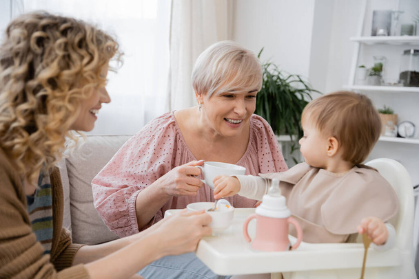 cheerful mature woman holding tea cup and looking at granddaughter having breakfast in kitchen - Foto, Bild