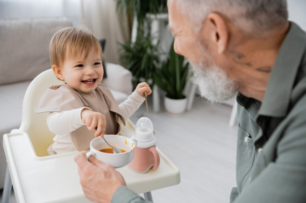cheerful baby holding spoon near bowl with puree and bearded grandfather on blurred foreground in kitchen - Photo, Image