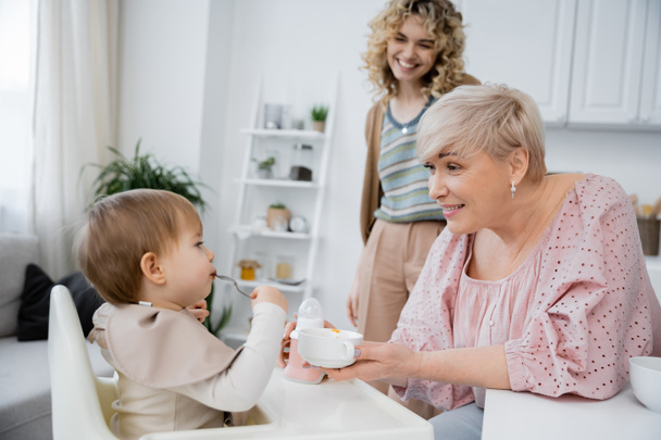 mulher de meia-idade feliz segurando tigela perto da neta da criança com colher durante o café da manhã na cozinha - Foto, Imagem