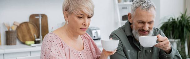 middle aged woman and smiling bearded man drinking tea in kitchen, banner - Foto, imagen