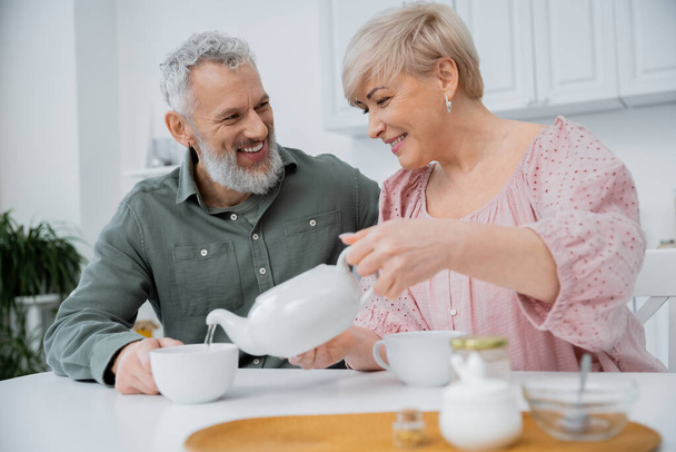 happy middle aged woman pouring tea near cheerful bearded husband in kitchen - Photo, Image
