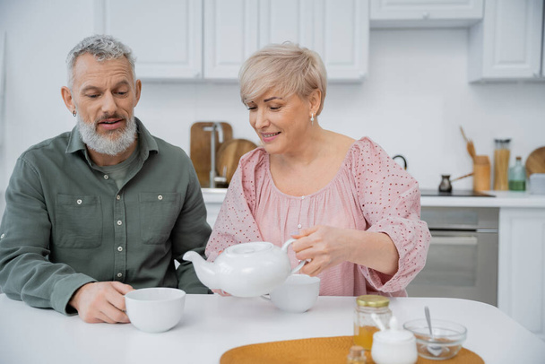 szczęśliwy w średnim wieku kobieta leje herbatę w pobliżu brodatego męża podczas śniadania w kuchni - Zdjęcie, obraz