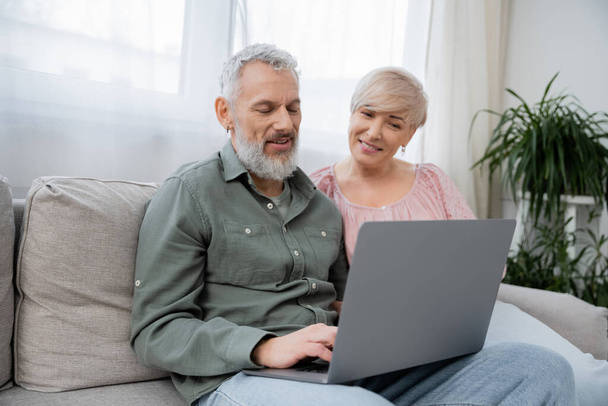 positive mature couple watching movie on laptop while sitting on couch in living room - Foto, Bild