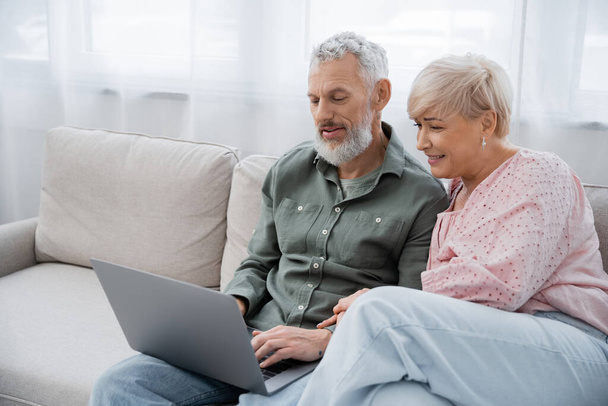 joyful middle aged woman and bearded man sitting on sofa in living room and watching movie on laptop - Fotografie, Obrázek