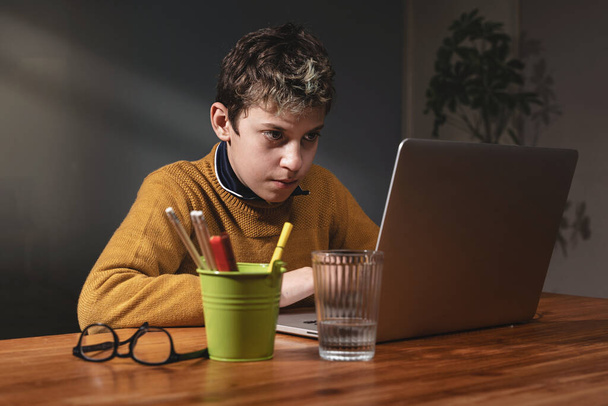 Skupiony 10-latek korzystający z laptopa - W słabo oświetlonym pomieszczeniu chłopiec koncentruje się na ekranie laptopa ze światłem z okna. Szklanki, szklanki i uchwyt na długopis na drewnianym biurku. - Zdjęcie, obraz