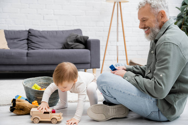 bearded man smiling near toddler granddaughter playing with toy car near building blocks on floor in living room - Fotó, kép