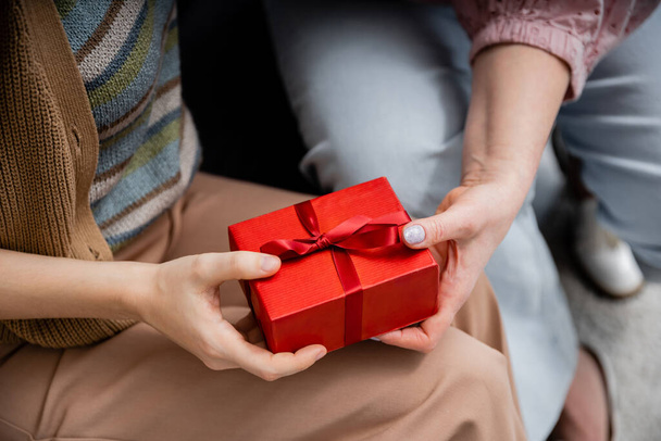 partial view of woman holding red gift box near mature mother while sitting at home - Foto, Imagen