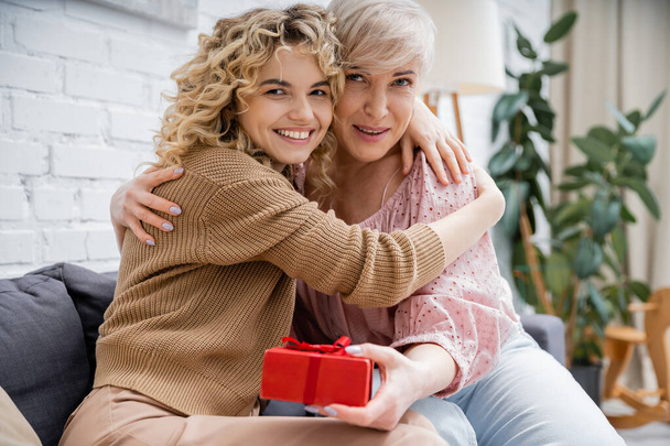 gelukkig volwassen vrouw met volwassen dochter omarmen en kijken naar camera in de buurt geschenk doos in woonkamer - Foto, afbeelding