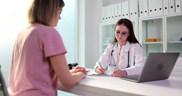 Jonge arts in bril schrijft medicijnen voor voor voor patiënten die aan tafel zitten in de buurt van een open laptop. Gericht specialist op afspraak met vrouw slow motion - Video