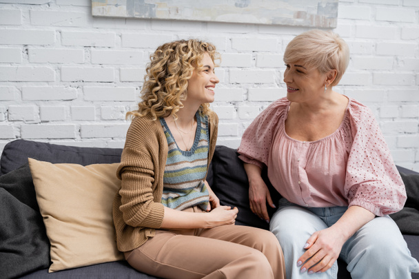 cheerful woman with blonde wavy hair talking to smiling middle aged mother on sofa in living room - Photo, image