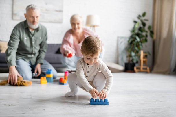 baby girl playing with building blocks on floor in living room near grandparents on blurred background - 写真・画像