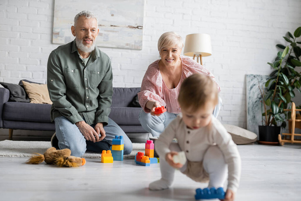 gelukkige middelbare leeftijd grootouders zitten op de vloer in de buurt van bouwstenen, terwijl klein meisje spelen met speelgoed auto in de woonkamer - Foto, afbeelding