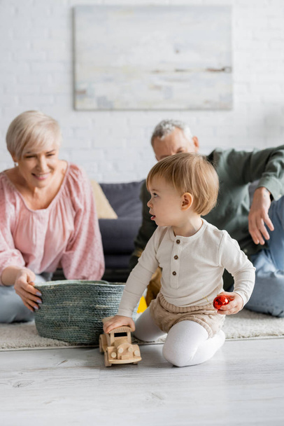little child playing with toy car near wicker basket and smiling grandparents on blurred background in living room - Photo, Image