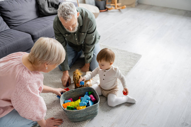 high angle view of mature couple and toddler girl near wicker basket with toys on floor in living room - Valokuva, kuva