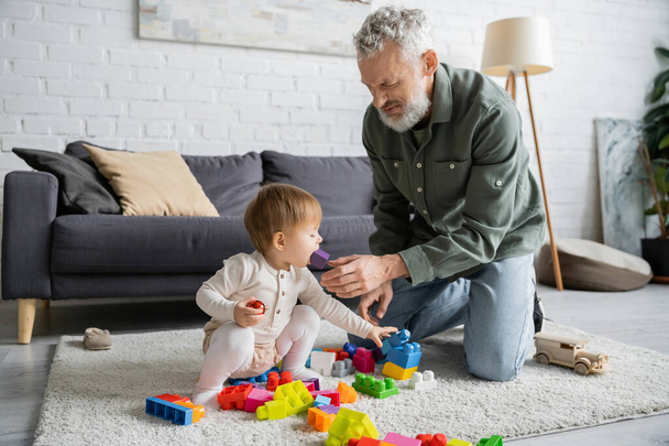 bearded man taking toy out of mouth of toddler granddaughter playing building blocks game on floor in living room - Fotó, kép