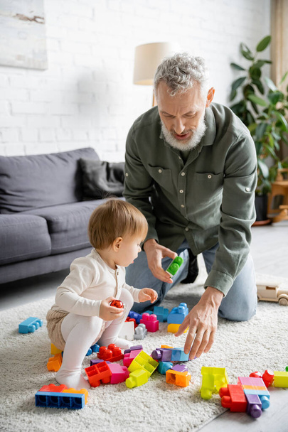 bearded man and little girl playing with multicolored building blocks on floor carpet in living room - Фото, изображение