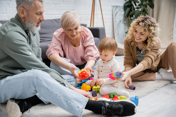 toddler girl playing with colorful building blocks near happy mother and grandparents on floor in living room - Foto, Bild