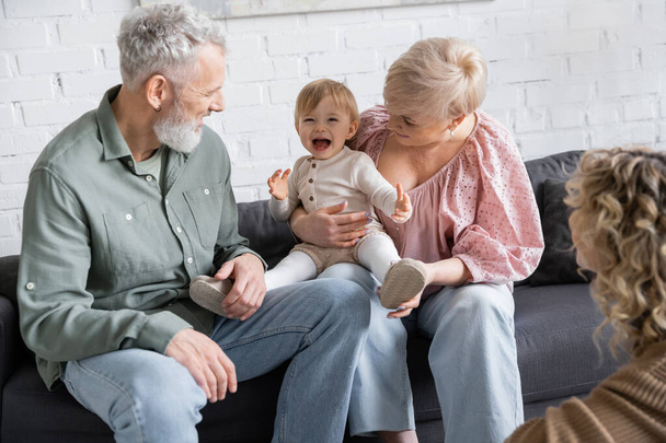 беззаботный ребенок смеется рядом с счастливыми бабушкой и дедушкой и размытой матерью в гостиной - Фото, изображение
