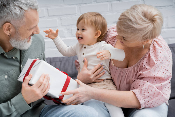 mature couple holding gift box near overjoyed granddaughter in living room - Photo, Image