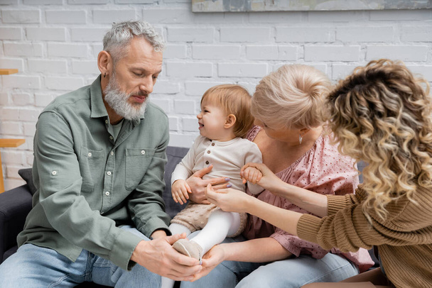 mature couple touching leg of cheerful granddaughter while sitting on sofa in living room - Photo, Image