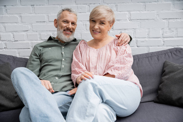 joyful middle aged couple sitting on sofa in living room and smiling at camera - Фото, изображение