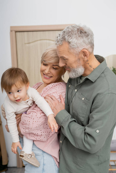 joyful woman embracing little granddaughter near bearded husband in hall at home - Photo, Image