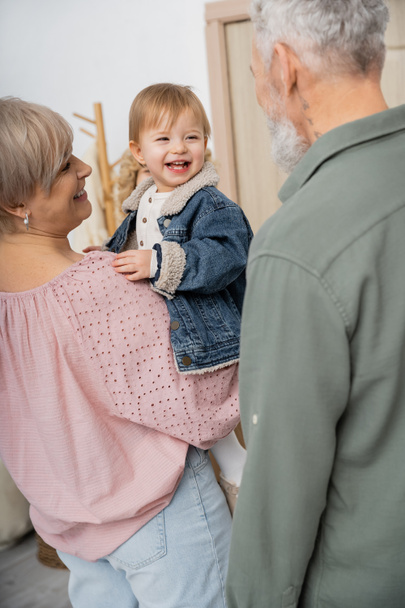 middle aged woman holding cheerful granddaughter in denim jacket near family at home - Photo, Image