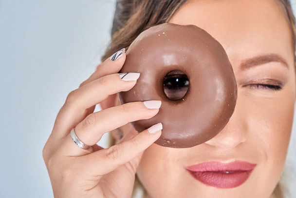 Vista de perto de uma mulher segurando um donut perto do rosto com suas unhas de manicure. Conceito de manicure e alimentação. - Foto, Imagem