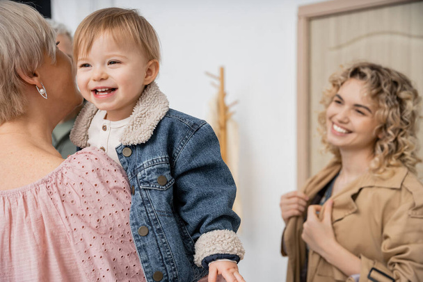 happy child in denim jacket smiling in hands of granny near blurred mother at door of apartment - Фото, изображение