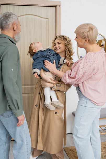 cheerful woman in trench coat holding little daughter in denim jacket while visiting mature parents at home - Foto, Bild