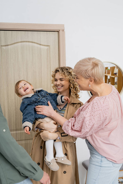 cheerful woman holding toddler daughter in denim jacket near while visiting happy parents at home - Foto, afbeelding