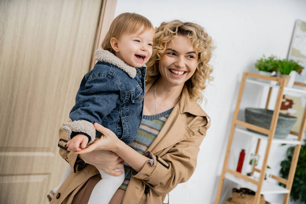 joyful blonde woman with wavy hair holding carefree daughter in denim jacket near entrance door at home - Foto, afbeelding